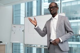 Young confident male teacher explaining audience financial information while standing by whiteboard during presentation
